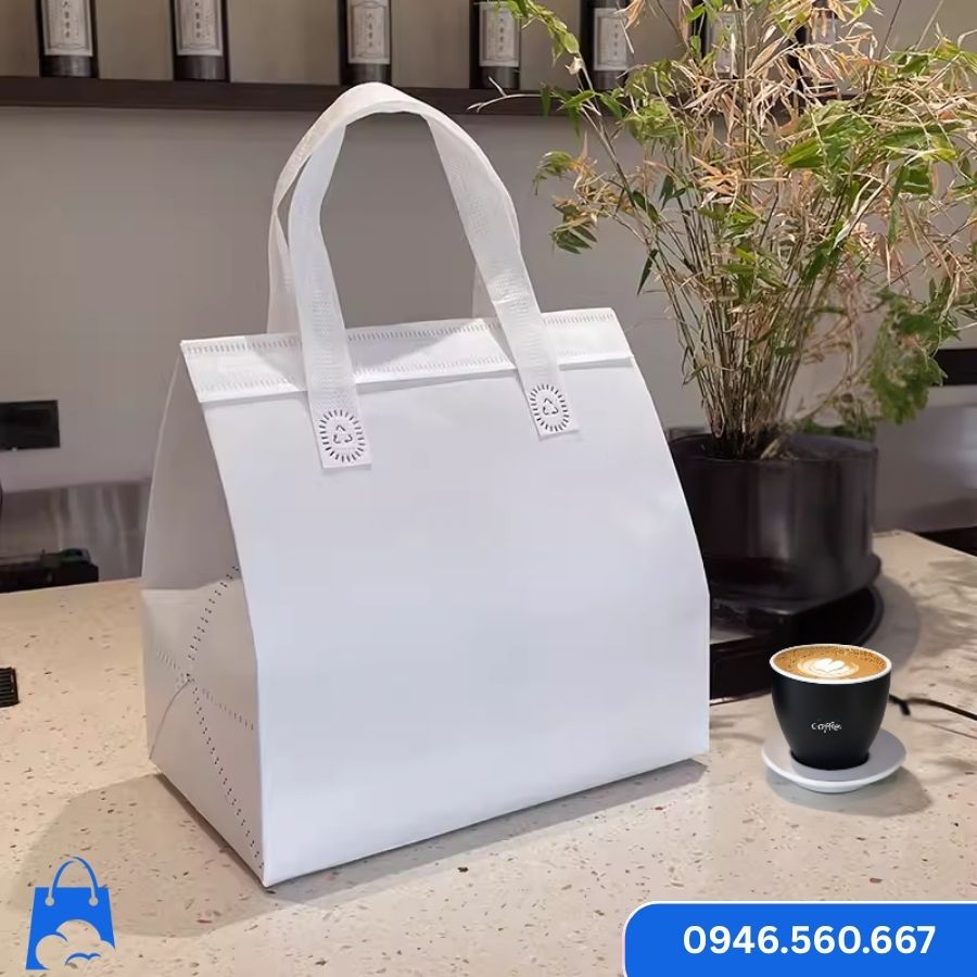 Túi giữ nhiệt trắng trơn đặt cạnh ly cà phê, kiểu dáng đơn giản, quai xách trắng đồng bộ, thích hợp cho quán cà phê, trà sữa take-away cần in logo riêng.